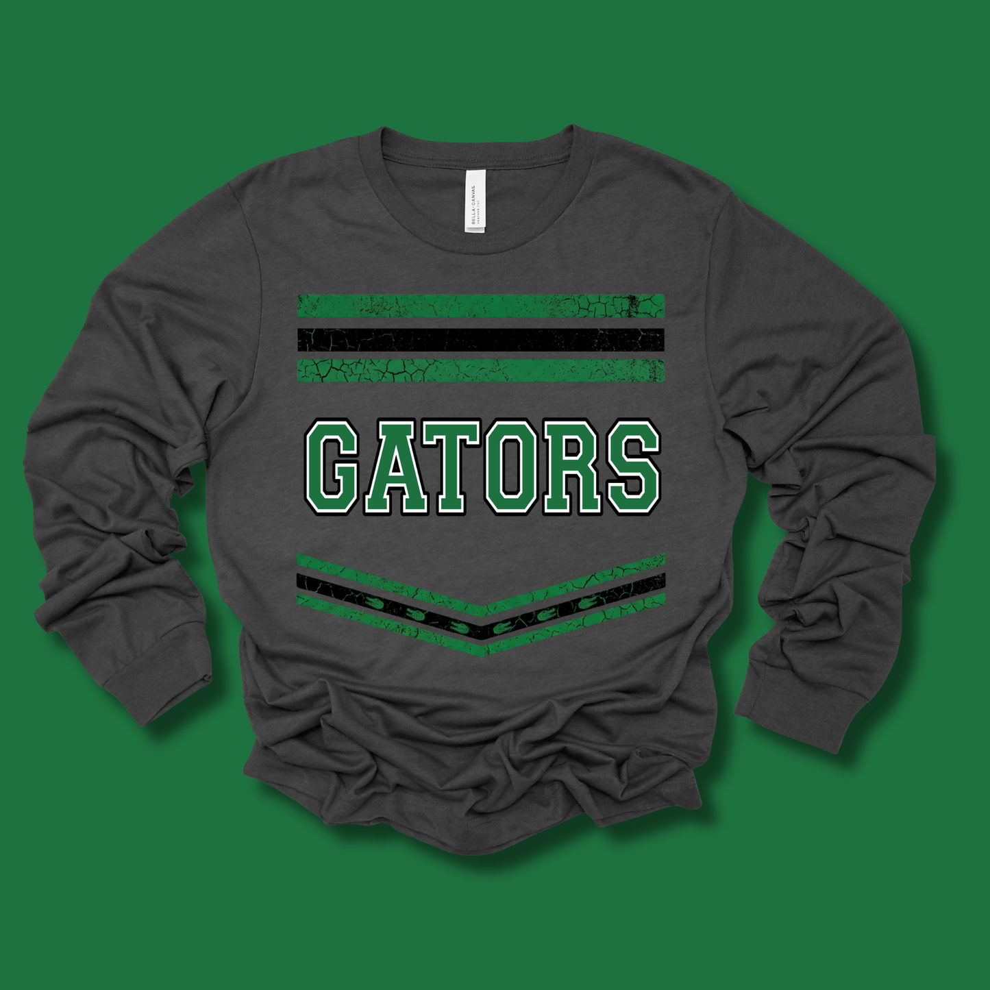 A dark gray long-sleeve t-shirt with 'GATORS' printed in green varsity letters across the chest, flanked by green and black horizontal stripes, laid flat on a solid green background.
