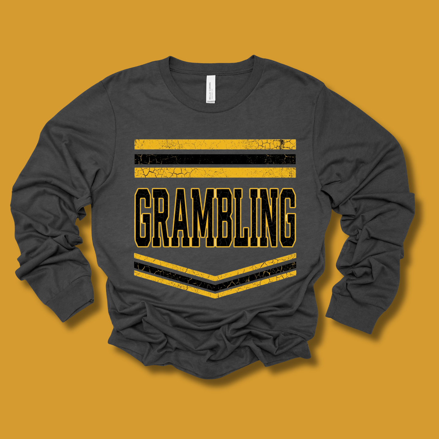 A dark charcoal gray long-sleeve t-shirt with a distressed yellow and black graphic reading 'GRAMBLING' on the front, displayed against a solid mustard yellow background.