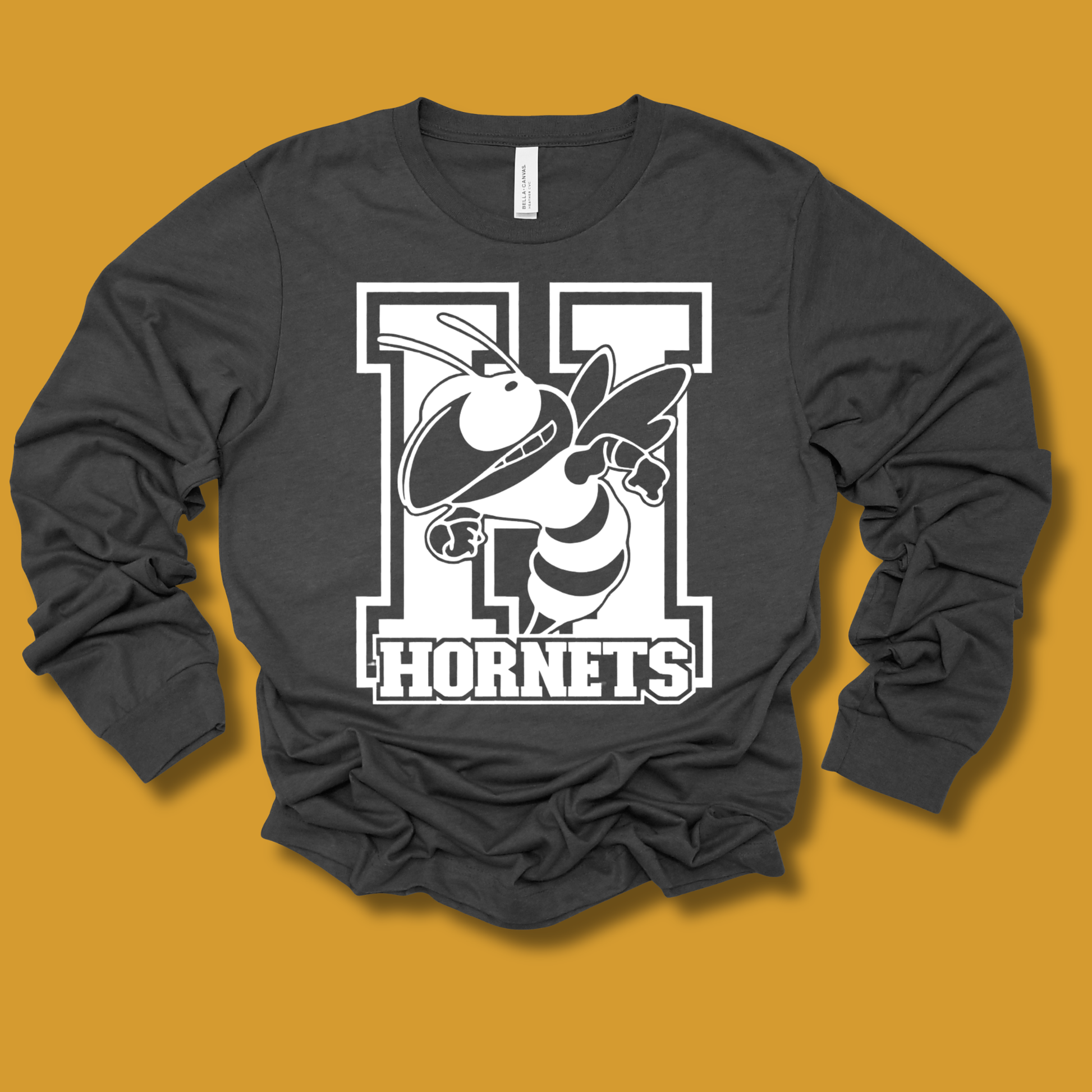 A dark charcoal gray long-sleeve t-shirt with a large white and black 'H Hornets' graphic on the front, displayed flat against a solid mustard yellow background.