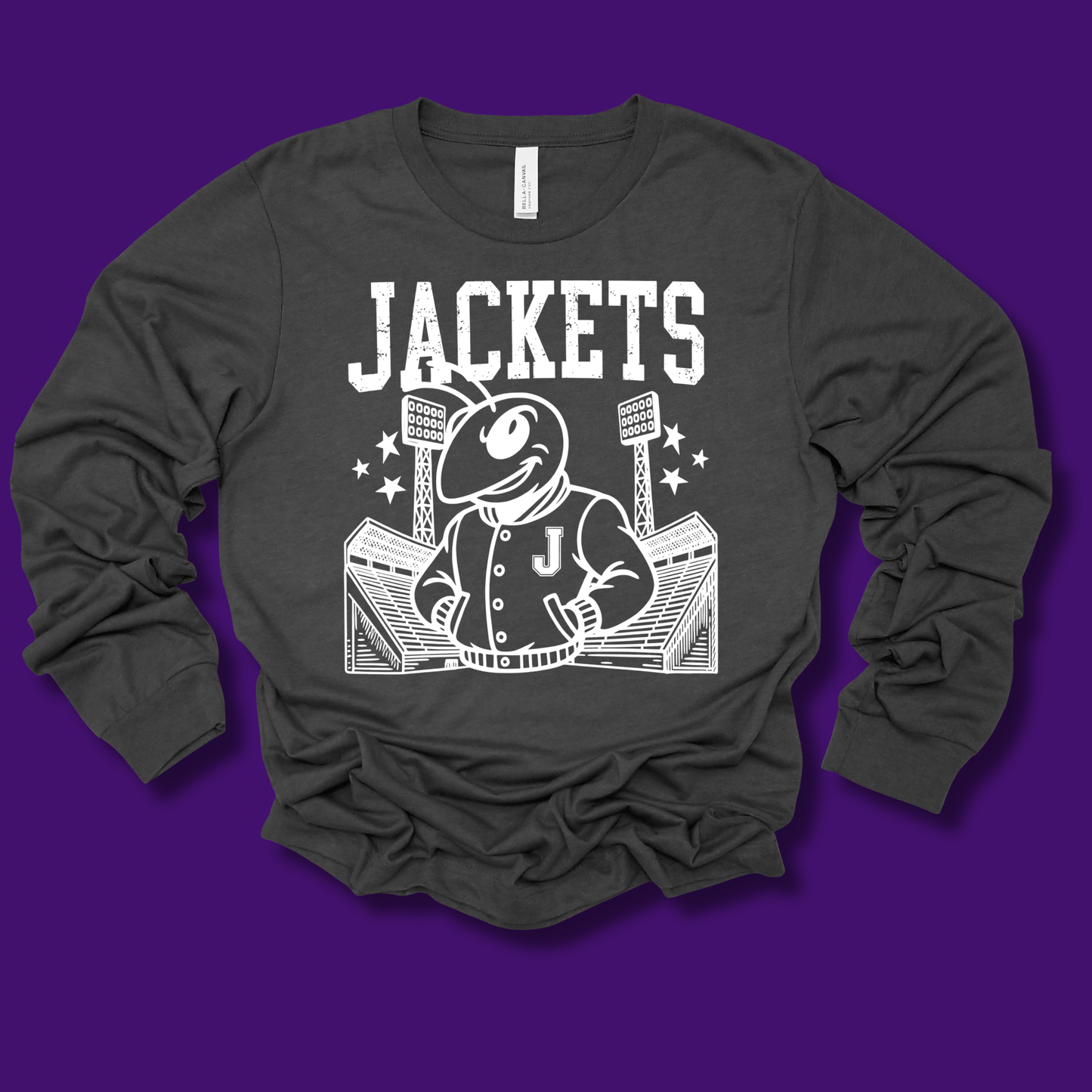 A dark charcoal gray long-sleeve t-shirt with a white graphic print of a mascot wearing a jacket and the word 'JACKETS' above it, laid flat on a solid purple background.