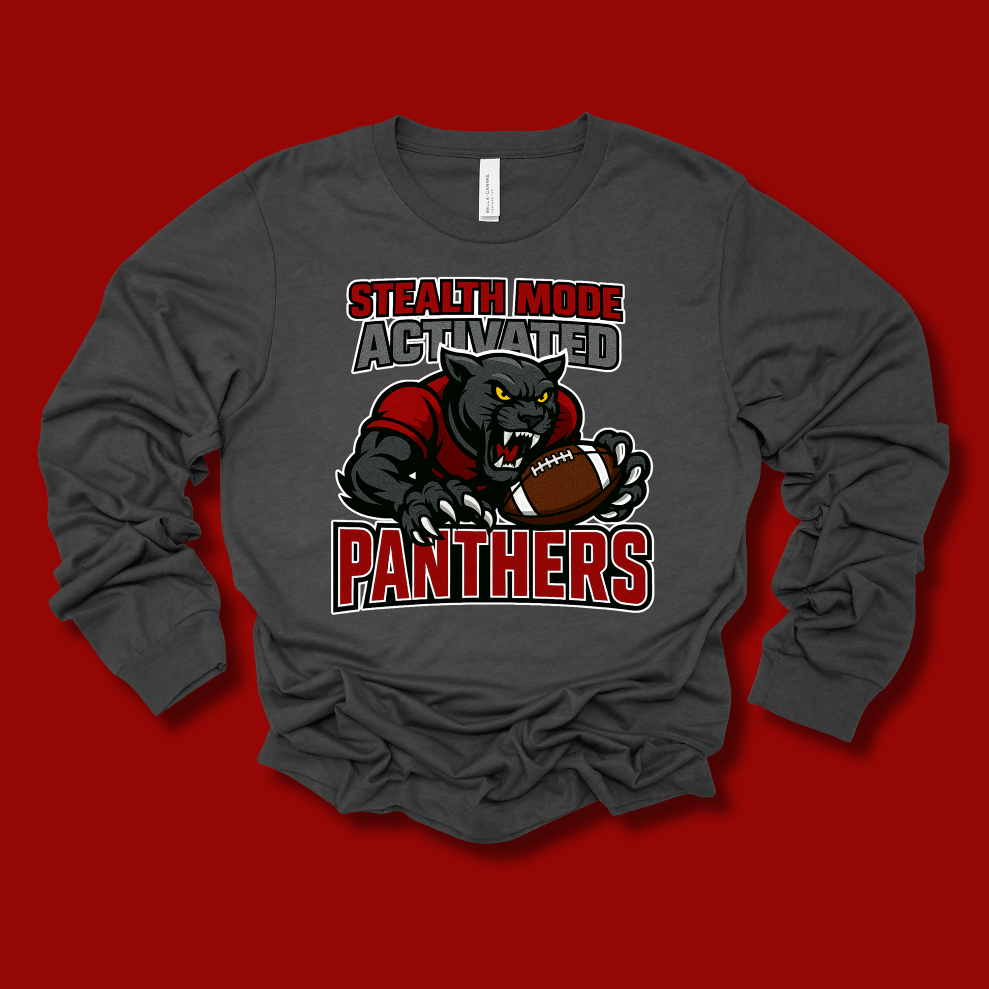 A dark gray long-sleeve t-shirt with a graphic of a black panther holding a football, with the text 'STEALTH MODE ACTIVATED' above and 'PANTHERS' below in red and white. The shirt is laid flat on a solid red background.