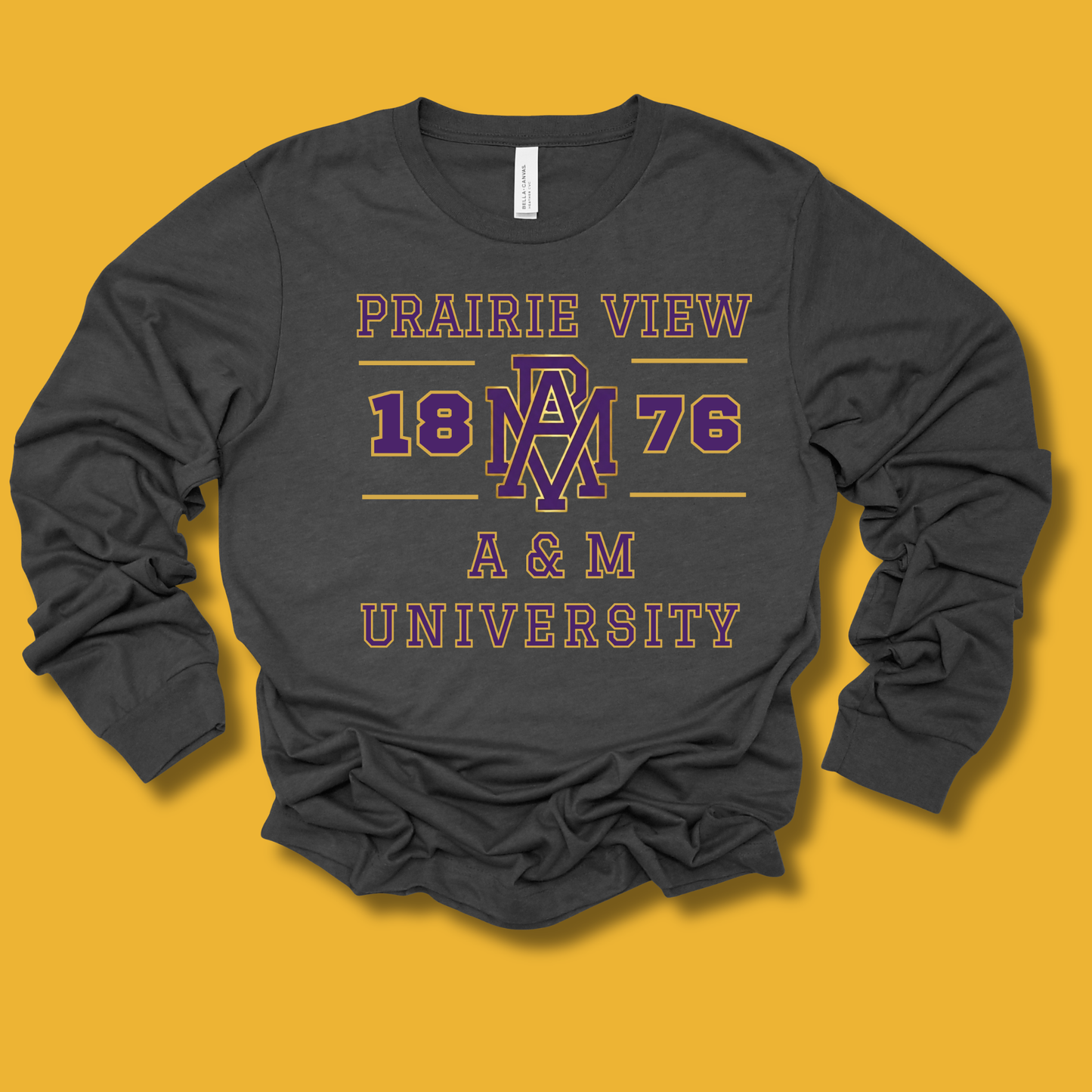 A dark charcoal gray long-sleeve t-shirt with a vintage-style graphic print reading 'PRAIRIE VIEW 1876 A & M UNIVERSITY' in purple and pink, laid flat on a solid mustard yellow background.