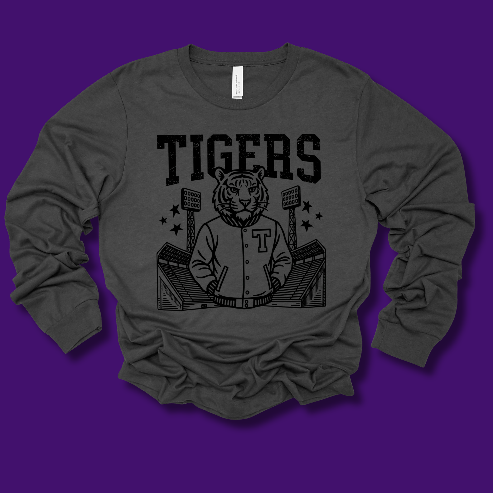 A dark charcoal gray long-sleeve t-shirt with a black graphic print of a tiger mascot in a baseball jersey and the word 'TIGERS' above it, displayed against a solid purple background.