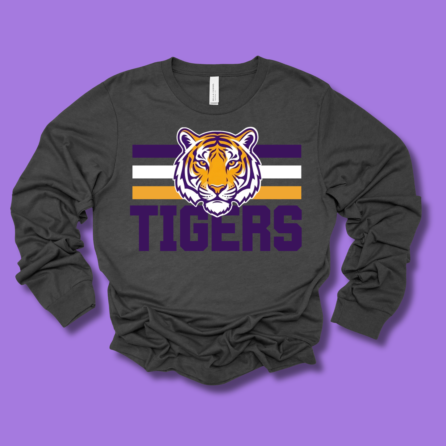 A dark charcoal gray long-sleeve t-shirt with a graphic of a tiger head over horizontal purple, white, and gold stripes, with the word 'TIGERS' printed below in purple. The shirt is displayed flat against a solid purple background.