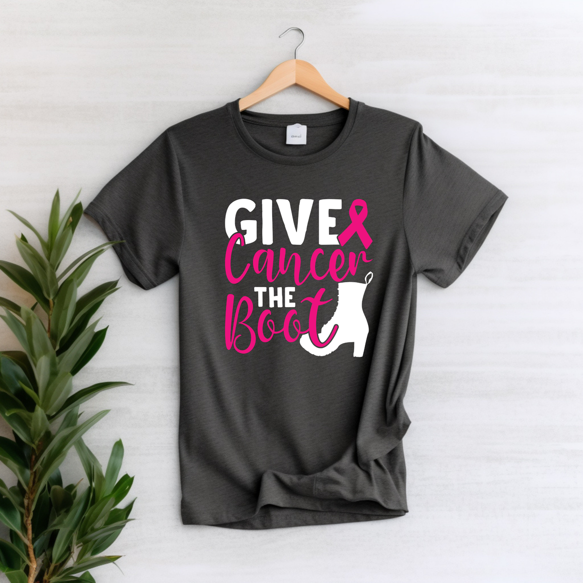 A dark charcoal gray t-shirt hanging on a wooden hanger against a light gray wood-paneled wall, featuring a graphic that reads 'GIVE Cancer THE Boot' in white and pink lettering with a white Christmas stocking silhouette. A green potted plant is visible on the left side of the frame.