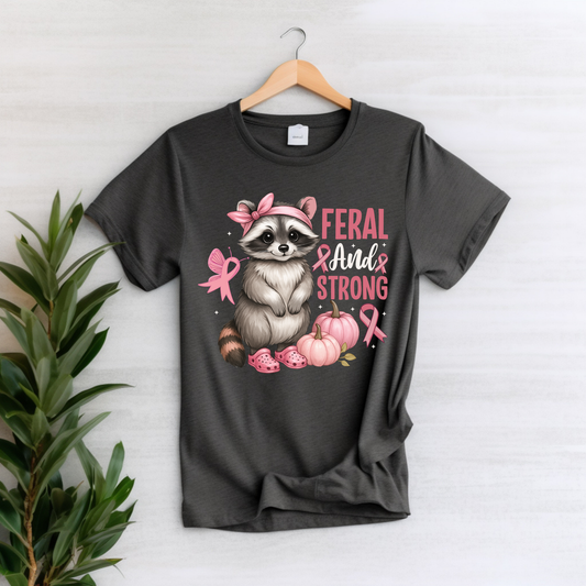 A dark charcoal gray graphic t-shirt hanging on a wooden hanger against a light wood paneled wall, with a green plant leaf on the left. The t-shirt features a graphic of a raccoon wearing a pink bow and headband, surrounded by pink bows, pumpkins, and the text 'FERAL AND STRONG'.