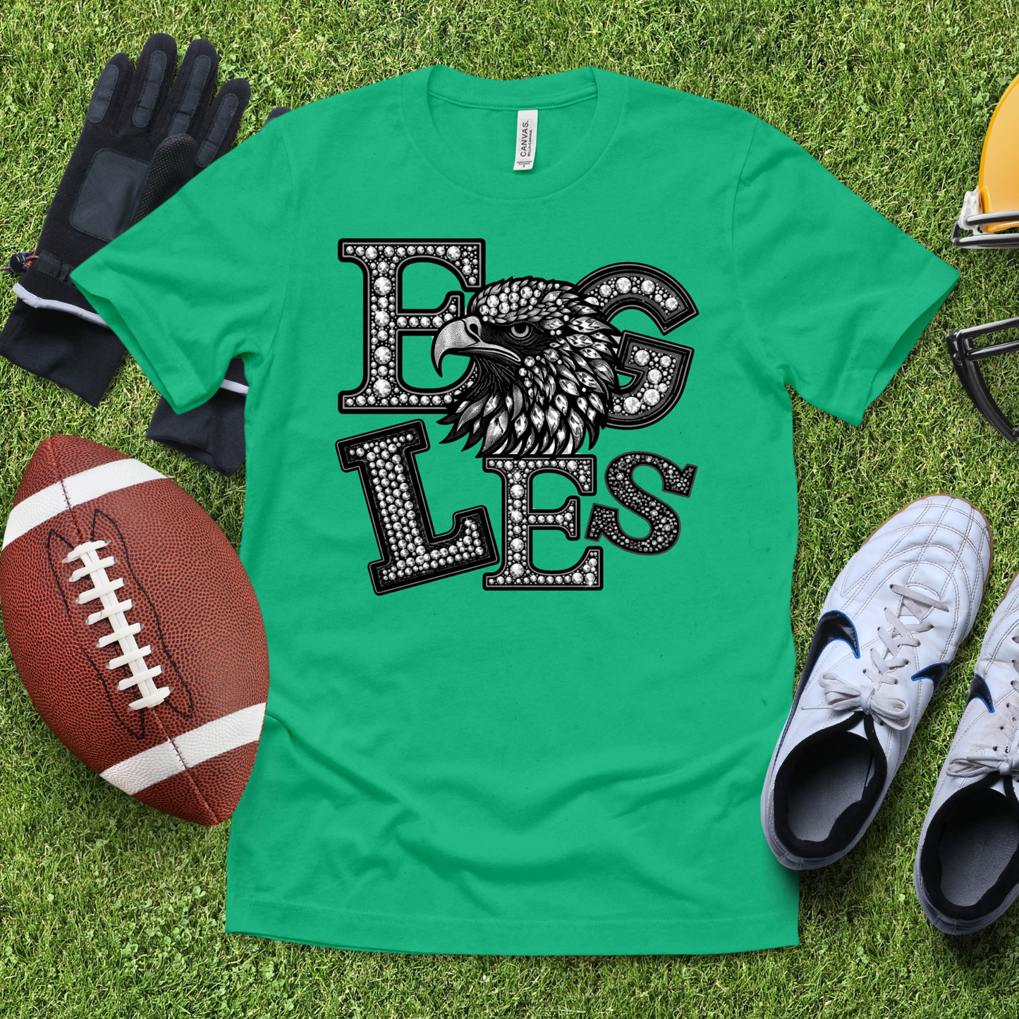 A green short-sleeve graphic T-shirt laid flat on grass, featuring a large black-and-white eagle head and the word 'EAGLES' in rhinestone-style letters. Surrounding props include a football, gloves, sneakers, and a helmet.