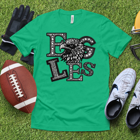 A green short-sleeve graphic T-shirt laid flat on grass, featuring a large black-and-white eagle head and the word 'EAGLES' in rhinestone-style letters. Surrounding props include a football, gloves, sneakers, and a helmet.