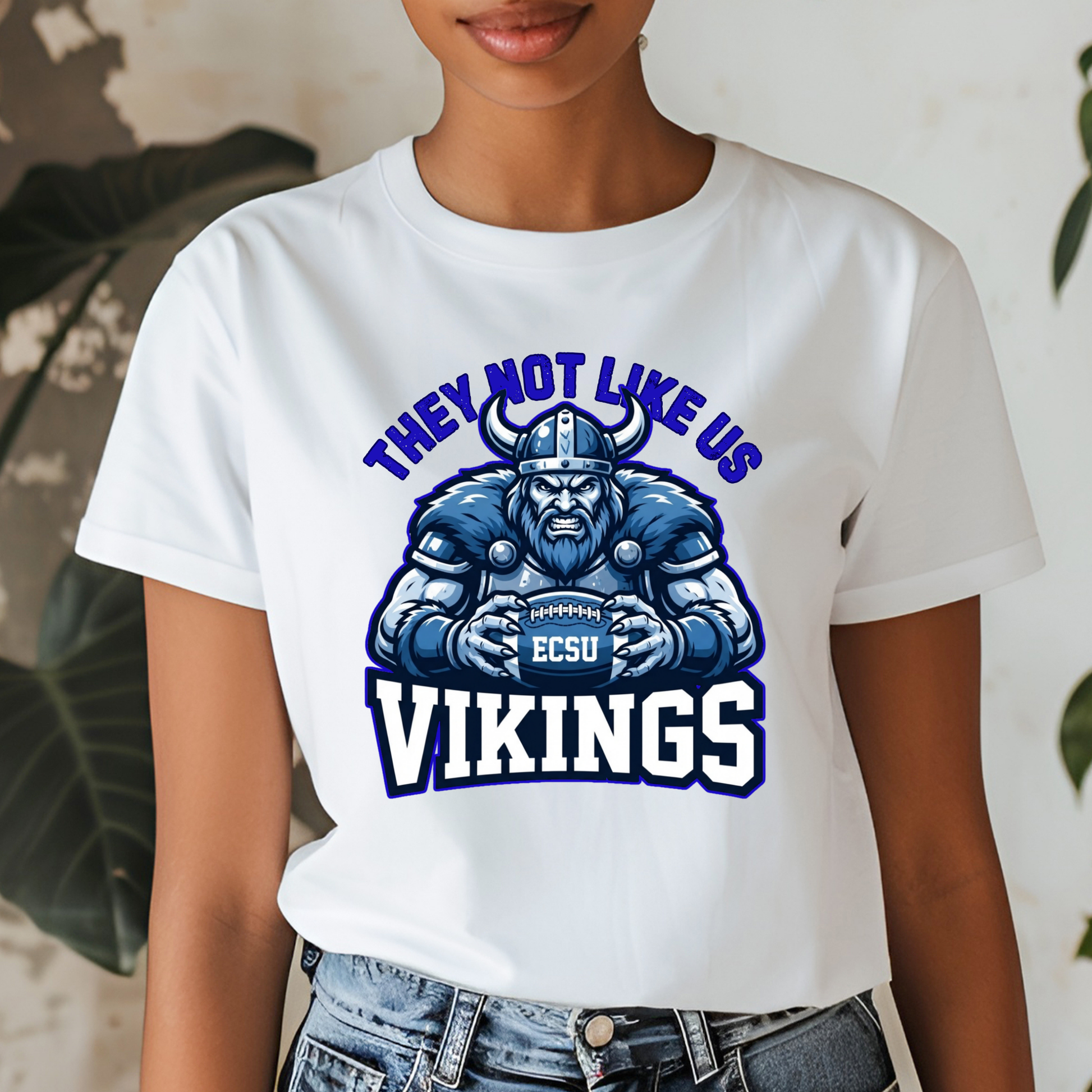 A woman wearing a white graphic t-shirt featuring a blue and purple illustration of a Viking football player with the text 'THEY NOT LIKE US VIKINGS' and 'ECSU' on the chest.