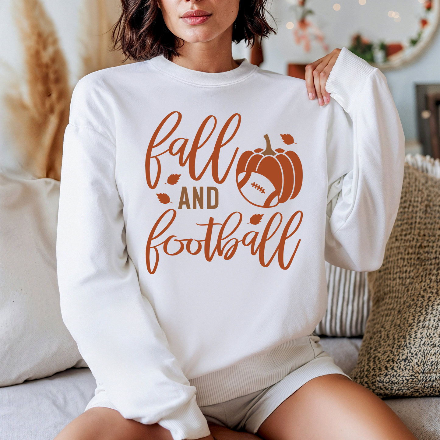 A woman wearing a white crewneck sweatshirt with a brown graphic that reads 'fall AND football' alongside a pumpkin and football design, sitting on a couch in a cozy indoor setting.