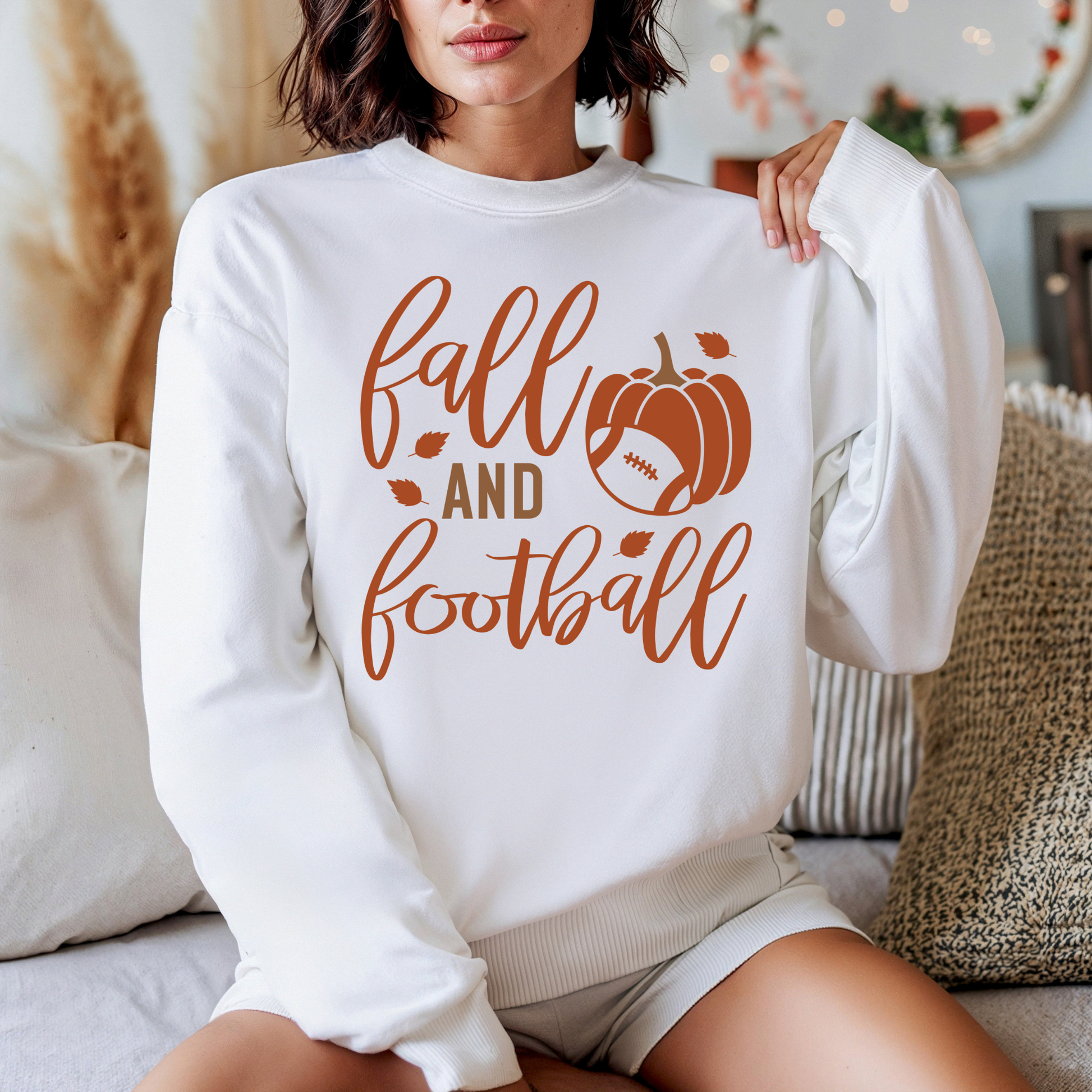 A woman wearing a white crewneck sweatshirt with a brown graphic that reads 'fall AND football' alongside a pumpkin and football design, sitting on a couch in a cozy indoor setting.