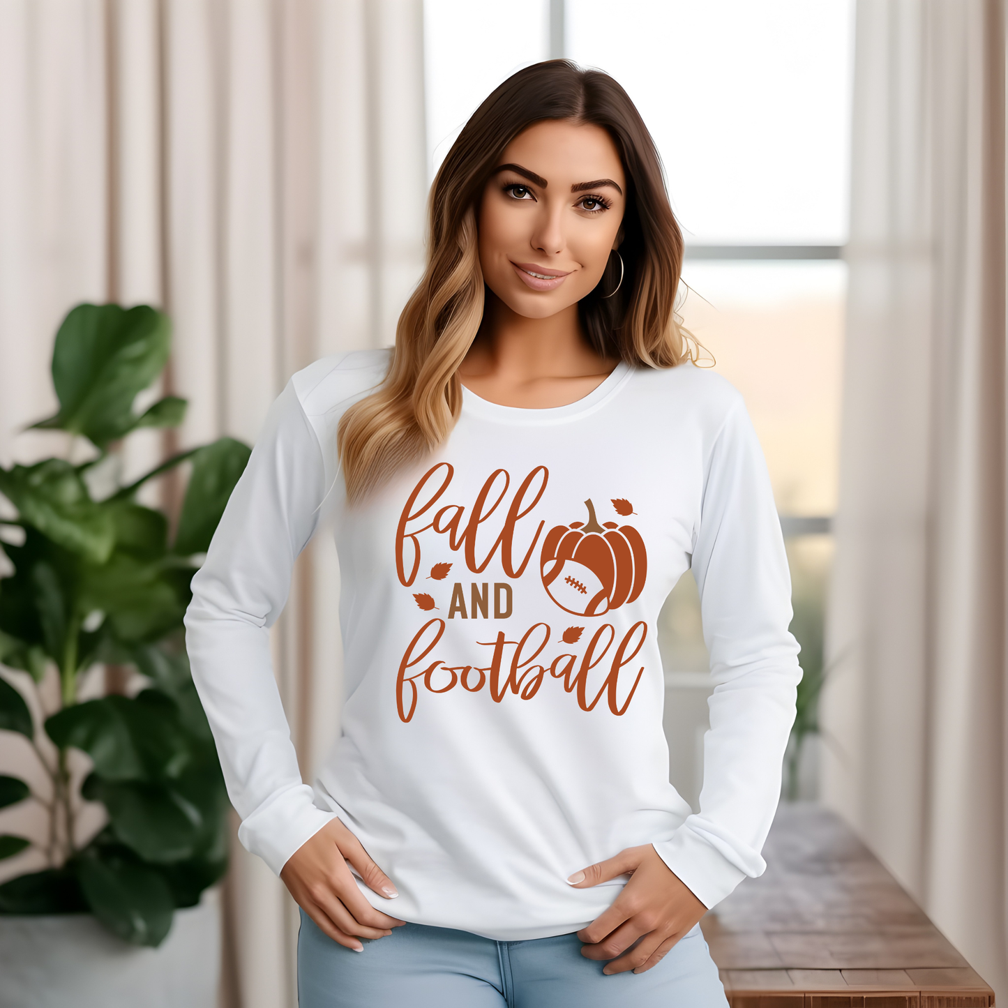 A woman wearing a white long-sleeve t-shirt with a brown 'fall and football' graphic, standing in a brightly lit room with curtains and a plant.
