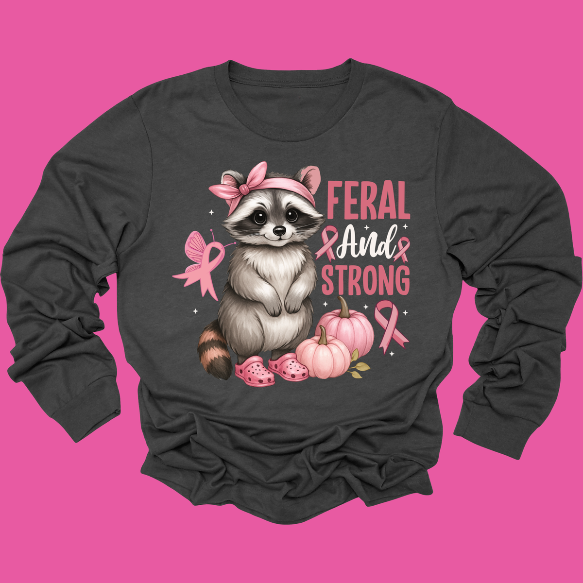 A dark charcoal gray long-sleeve t-shirt with a graphic of a cute raccoon wearing a pink bow and pink slippers, next to pumpkins and pink breast cancer awareness ribbons, with the text 'FERAL AND STRONG'. The shirt is displayed flat against a solid bright pink background.