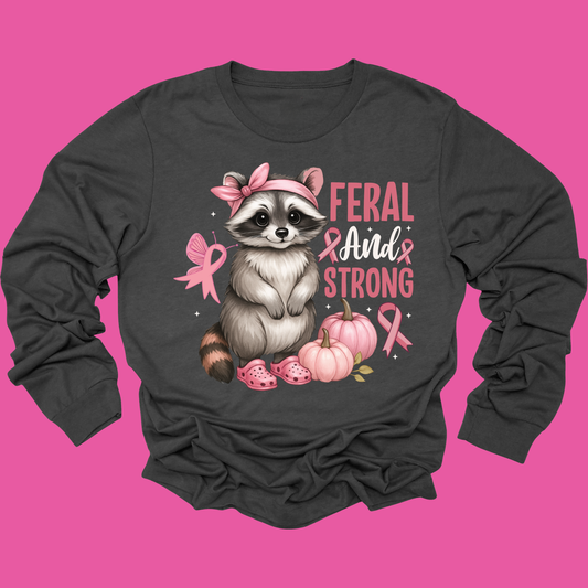 A dark charcoal gray long-sleeve t-shirt with a graphic of a cute raccoon wearing a pink bow and pink slippers, next to pumpkins and pink breast cancer awareness ribbons, with the text 'FERAL AND STRONG'. The shirt is displayed flat against a solid bright pink background.