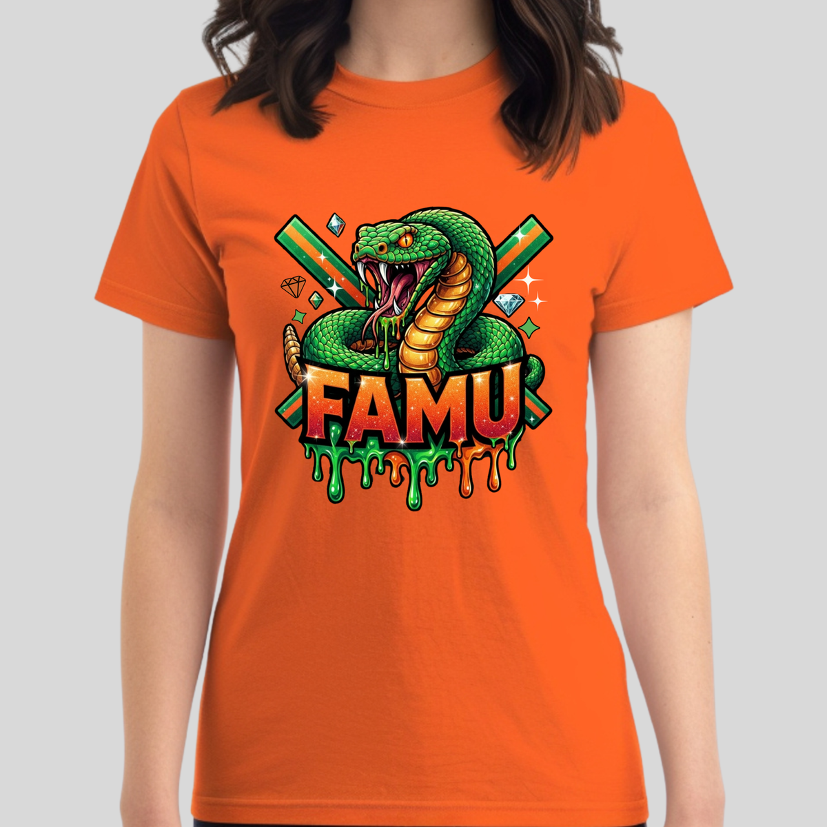 A woman wearing a bright orange short-sleeve t-shirt with a colorful graphic print on the front. The graphic features a green snake coiled around crossed green swords, diamonds, and the text 'FAMU' in red and orange with green dripping details, set against a solid gray background.