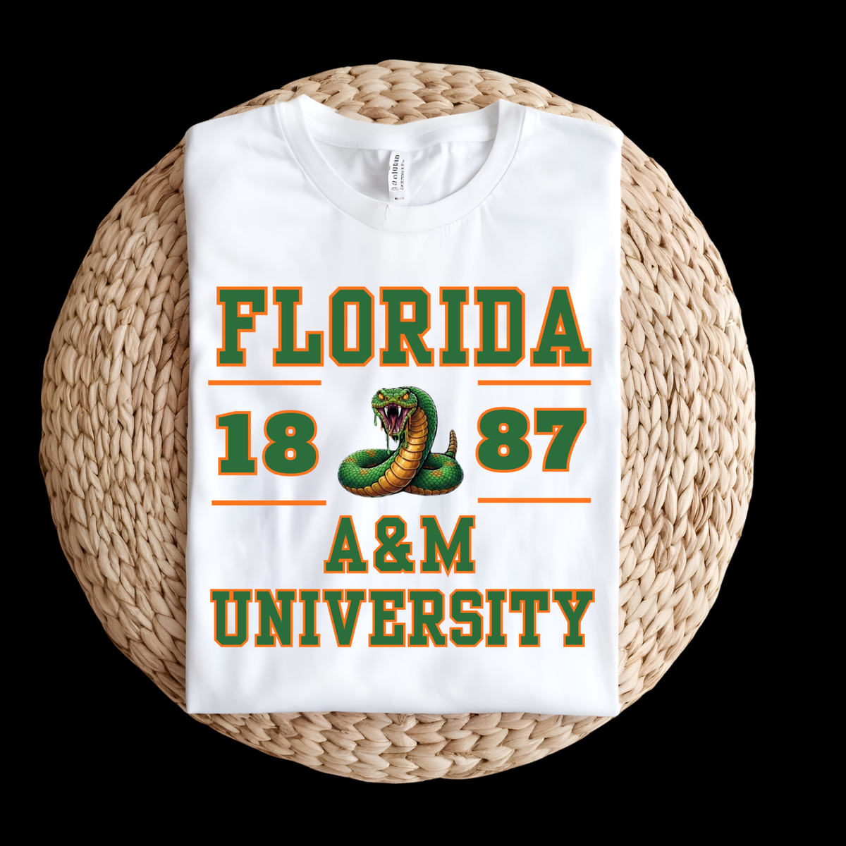 A folded white t-shirt with a green and orange 'Florida 1887 A&M University' graphic featuring a snake, displayed on a round woven straw mat against a black background.