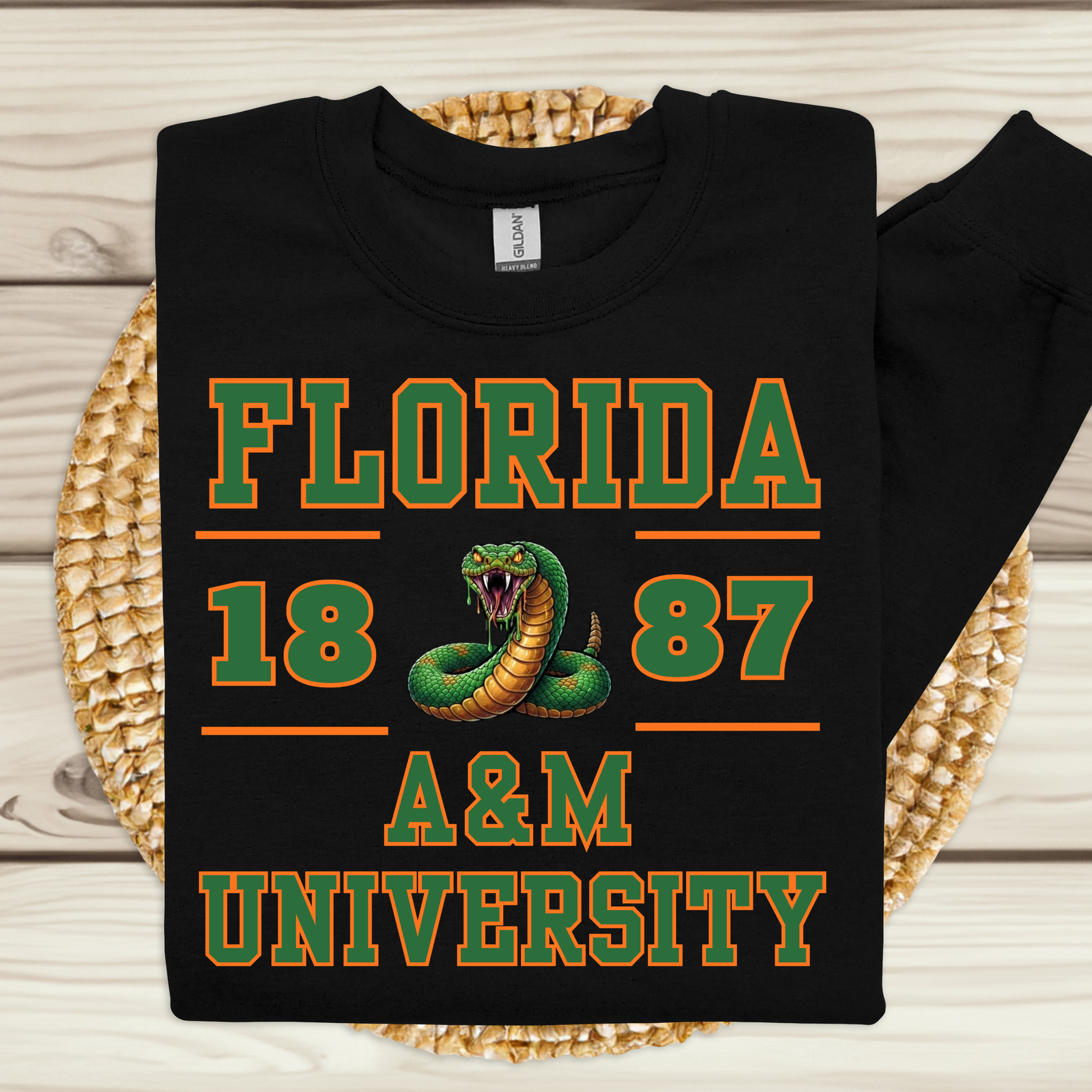 A black long-sleeve t-shirt with a green and orange graphic reading 'FLORIDA 1887 A&M UNIVERSITY' and featuring a snake illustration, laid flat on a woven straw mat over a light wood background.