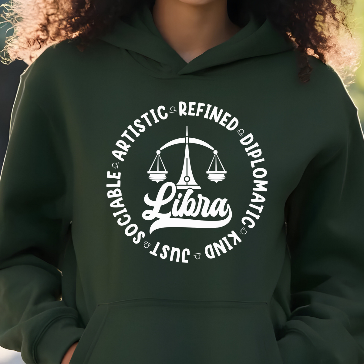 Woman wearing a white crewneck t-shirt featuring a black circular Libra zodiac graphic with a scales illustration, the word "Libra," and trait keywords like artistic, refined, diplomatic, kind, just, and sociable.