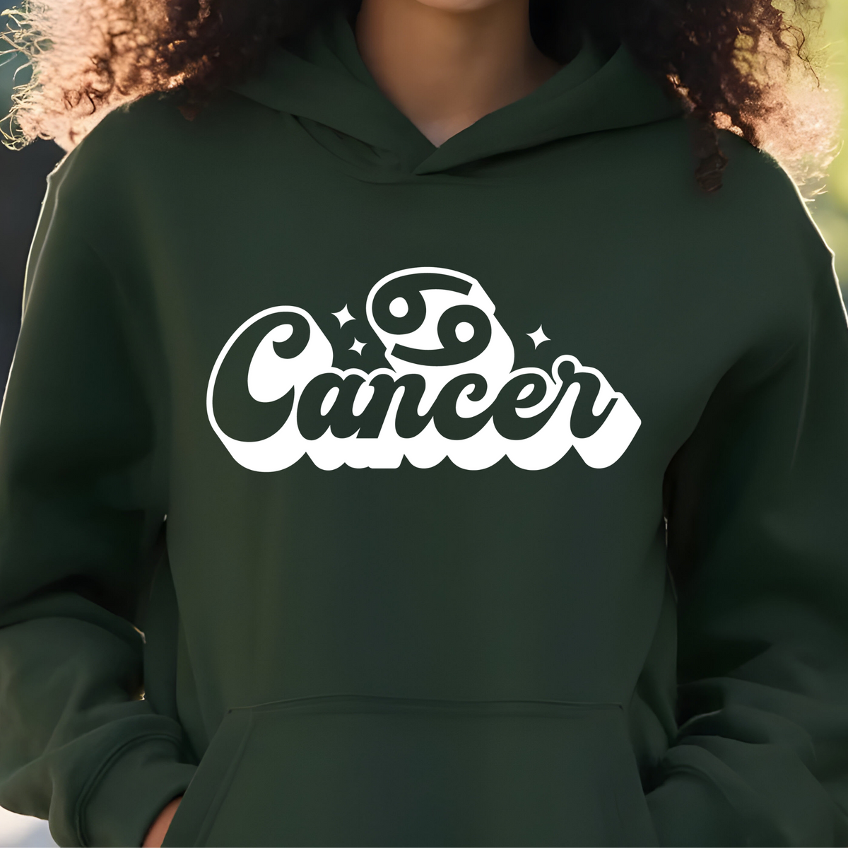 A person wearing a dark forest green hoodie with a white graphic print that reads 'Cancer' with the Cancer zodiac symbol. The photo is cropped from the neck down, showing the person's curly hair and hands in the hoodie pockets, set against a softly blurred outdoor background.