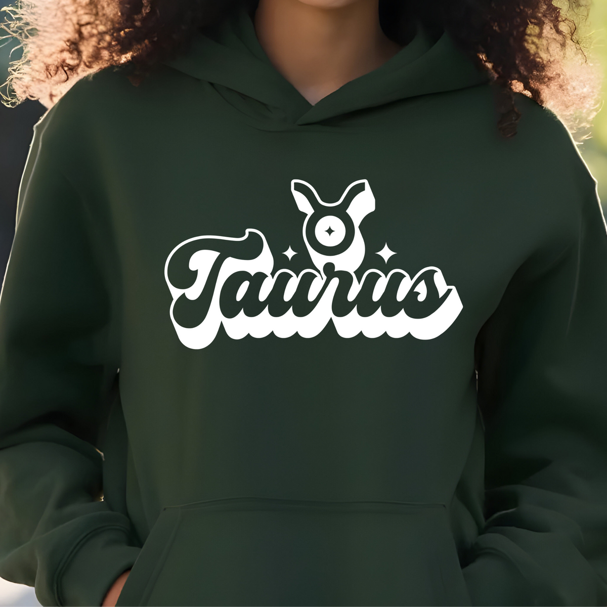 A person wearing a dark forest green hoodie with a white graphic print that reads 'Taurus' in stylized script and includes a Taurus zodiac symbol on the chest.