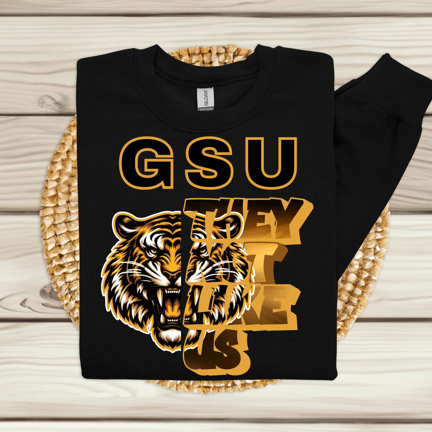 A black long-sleeve crewneck sweatshirt with a bold graphic print featuring the text 'GSU THEY'RE LEE US' in gold and a roaring tiger illustration, displayed flat on a woven placemat over a light wood background.