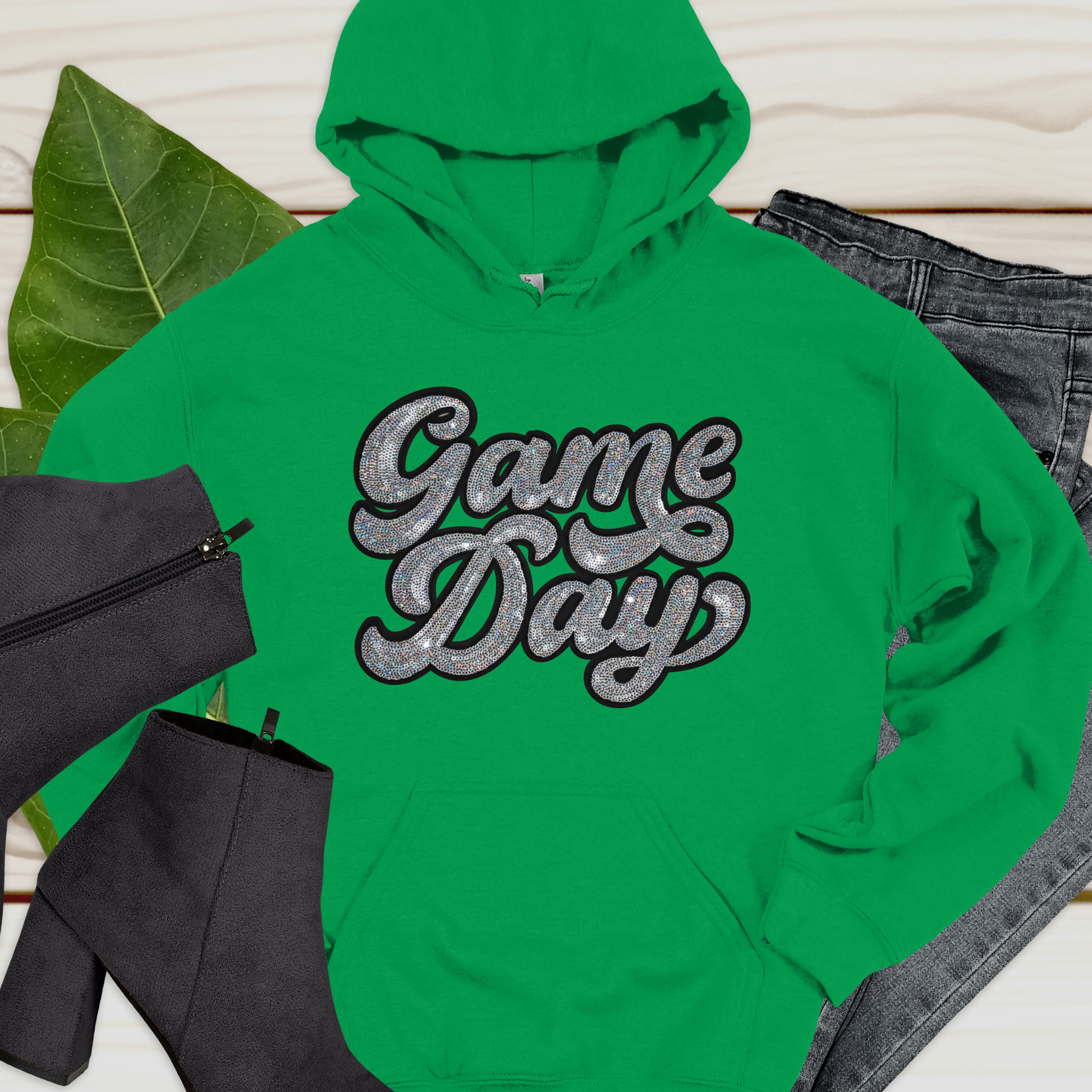 Green hoodie with 'Game Day' text on a light wooden surface with black bag and jeans.