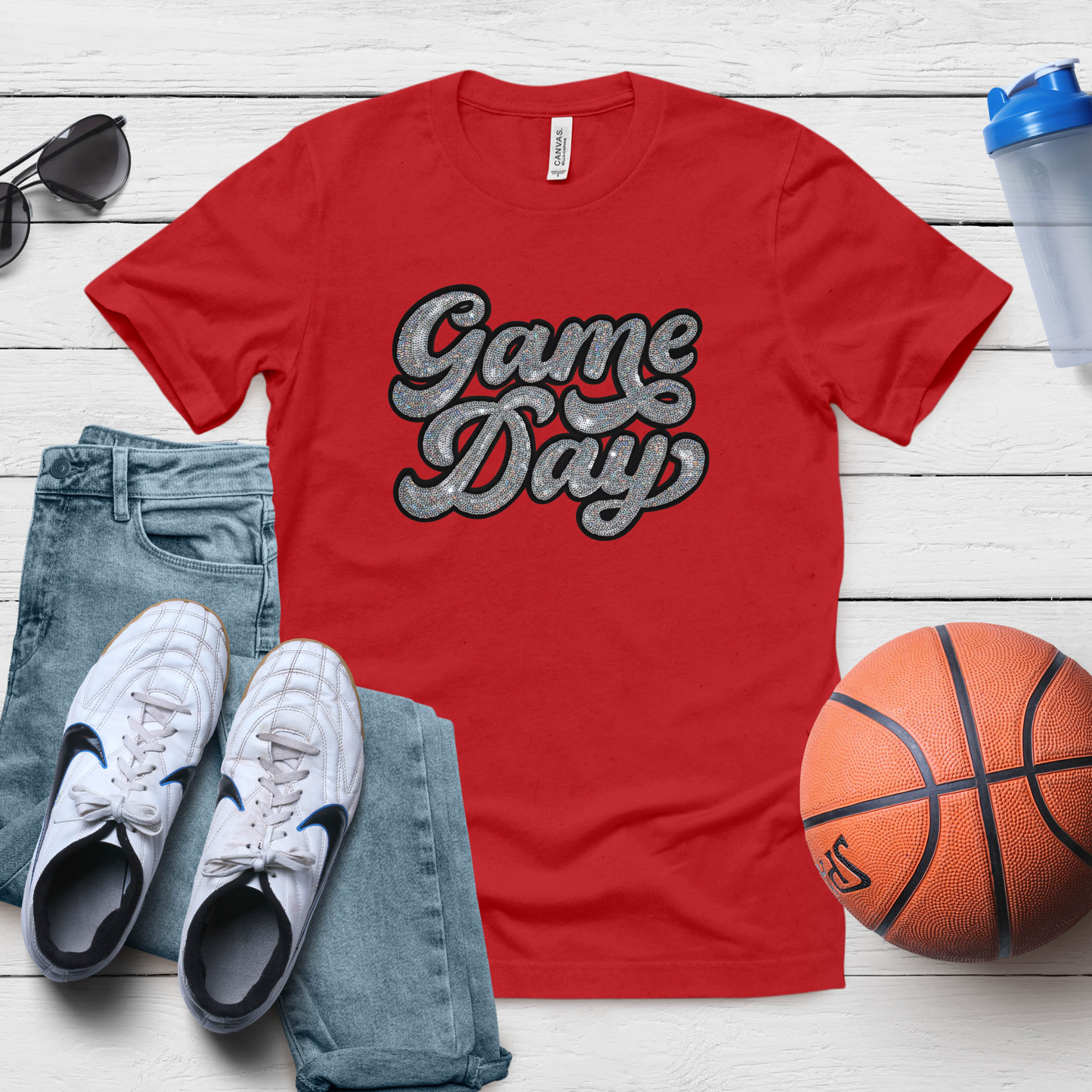 Red 'Game Day' t-shirt with white sneakers, blue jeans, a basketball, and sunglasses on a wooden surface.