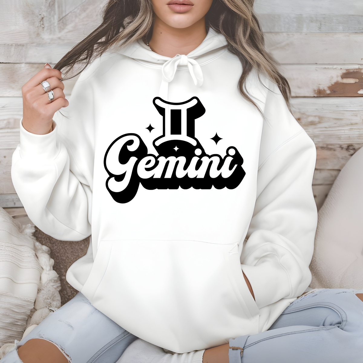 A woman wearing a white pullover hoodie with a black Gemini zodiac graphic on the chest, sitting against a rustic wood and beige cushion background.