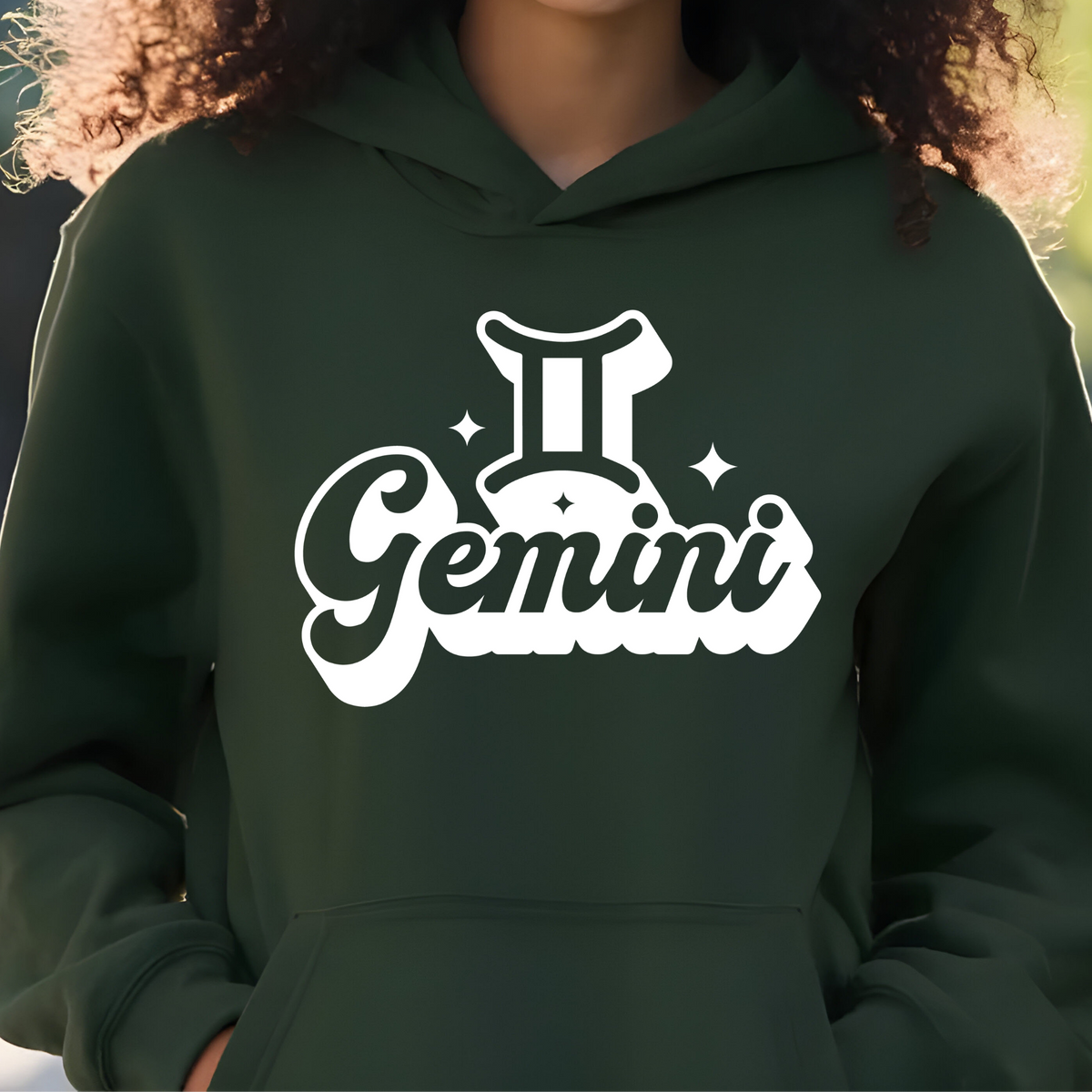 A person wearing a dark forest green hoodie with a white graphic print on the chest that reads 'Gemini' with a Gemini zodiac symbol. The image is cropped from the neck down, showing the person's curly hair and hands in the hoodie pockets.