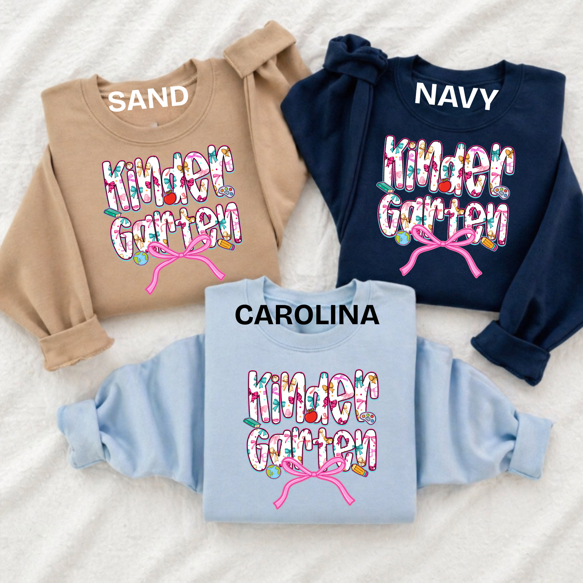 Three folded sweatshirts laid flat on a white textured fabric background, each with a colorful floral 'Kinder Garten' graphic and a pink bow design. The sweatshirts are in tan, navy, and light blue, with the names SAND, NAVY, and CAROLINA printed above the graphics.
