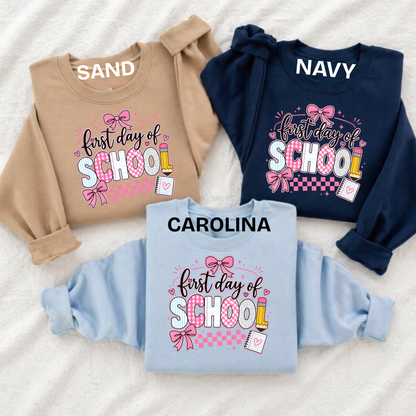Three long-sleeve sweatshirts laid flat on a white textured fabric background, each with a colorful 'BACK TO SCHOOL' graphic. The top-left sweatshirt is tan and labeled 'SAND', the top-right is navy and labeled 'NAVY', and the bottom-center is light blue and labeled 'CAROLINA'.