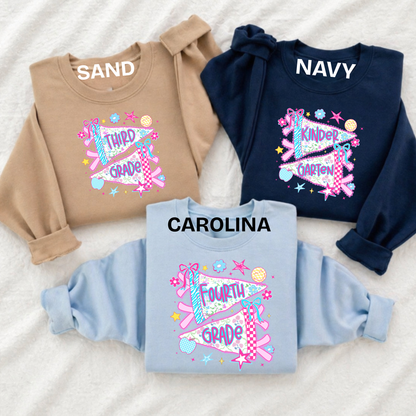 Three children's long-sleeve sweatshirts laid flat on a white textured fabric background, each with a large yellow bow and pencil graphic on the front and personalized names above the chest print: SAND, NAVY, and CAROLINA.