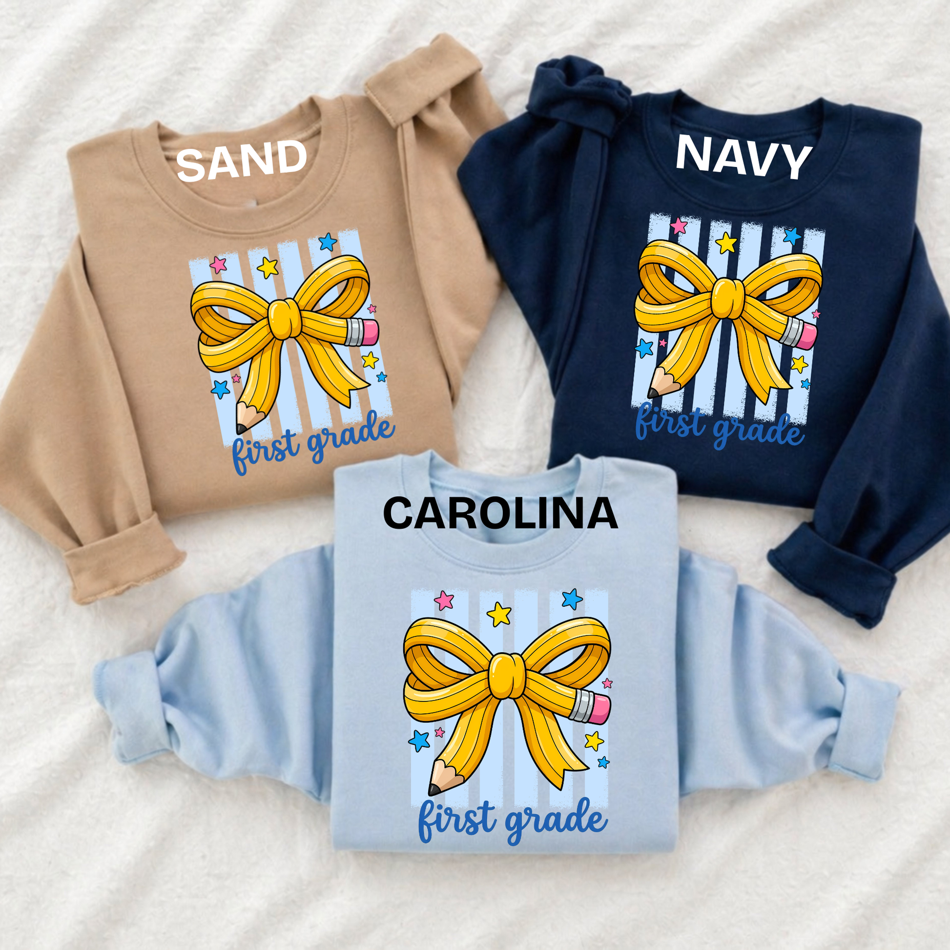 Three children's long-sleeve sweatshirts laid flat on a white textured fabric background, each with a large yellow bow and pencil graphic on the front and personalized names above the chest print: SAND, NAVY, and CAROLINA.