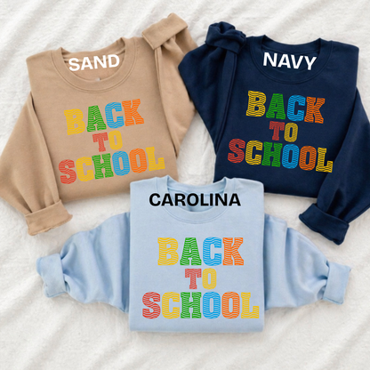 Three long-sleeve sweatshirts laid flat on a white textured fabric background, each with a colorful 'BACK TO SCHOOL' graphic. The top-left sweatshirt is tan and labeled 'SAND', the top-right is navy and labeled 'NAVY', and the bottom-center is light blue and labeled 'CAROLINA'.