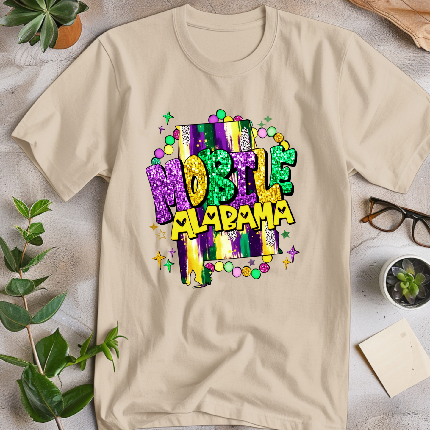Beige t-shirt with 'Moble Alabama' design on a light surface with plants and glasses.