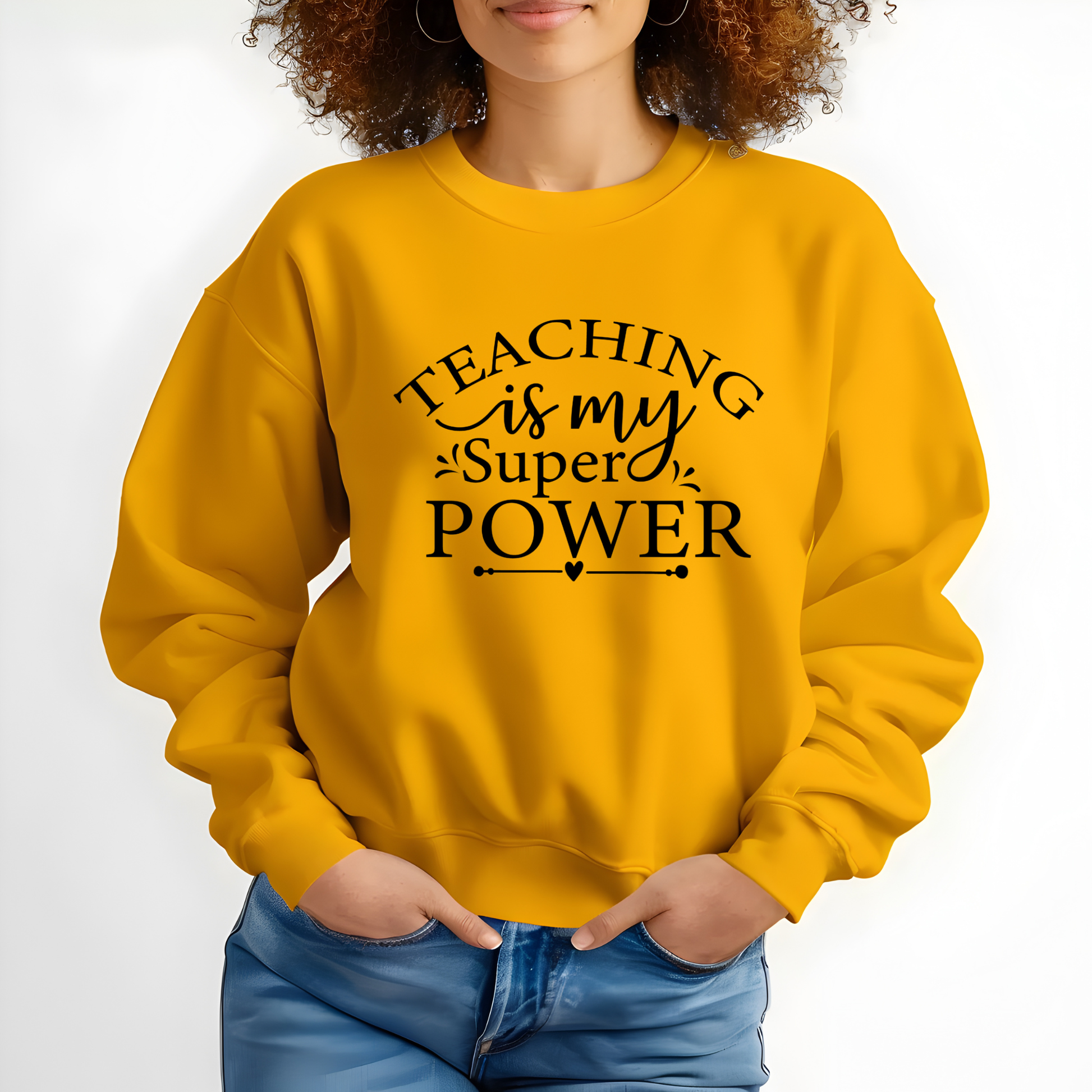 A woman wearing a mustard yellow crewneck sweatshirt with the black printed text 'TEACHING is my Super POWER' on the front, paired with blue jeans against a plain white background.