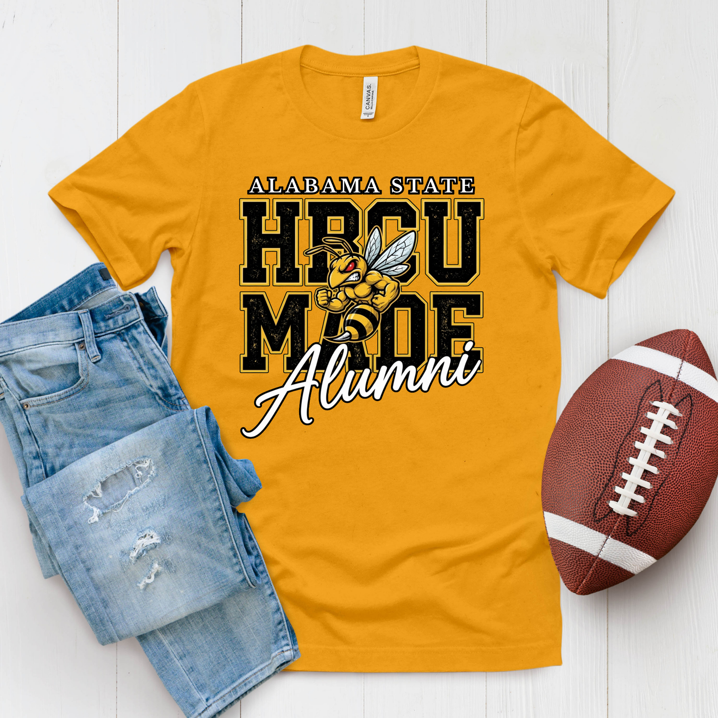 A gold crewneck t-shirt featuring a 'Hornets Nation' graphic with a hornet mascot, styled in a flat lay with blue jeans and a brown football on a white wood background.