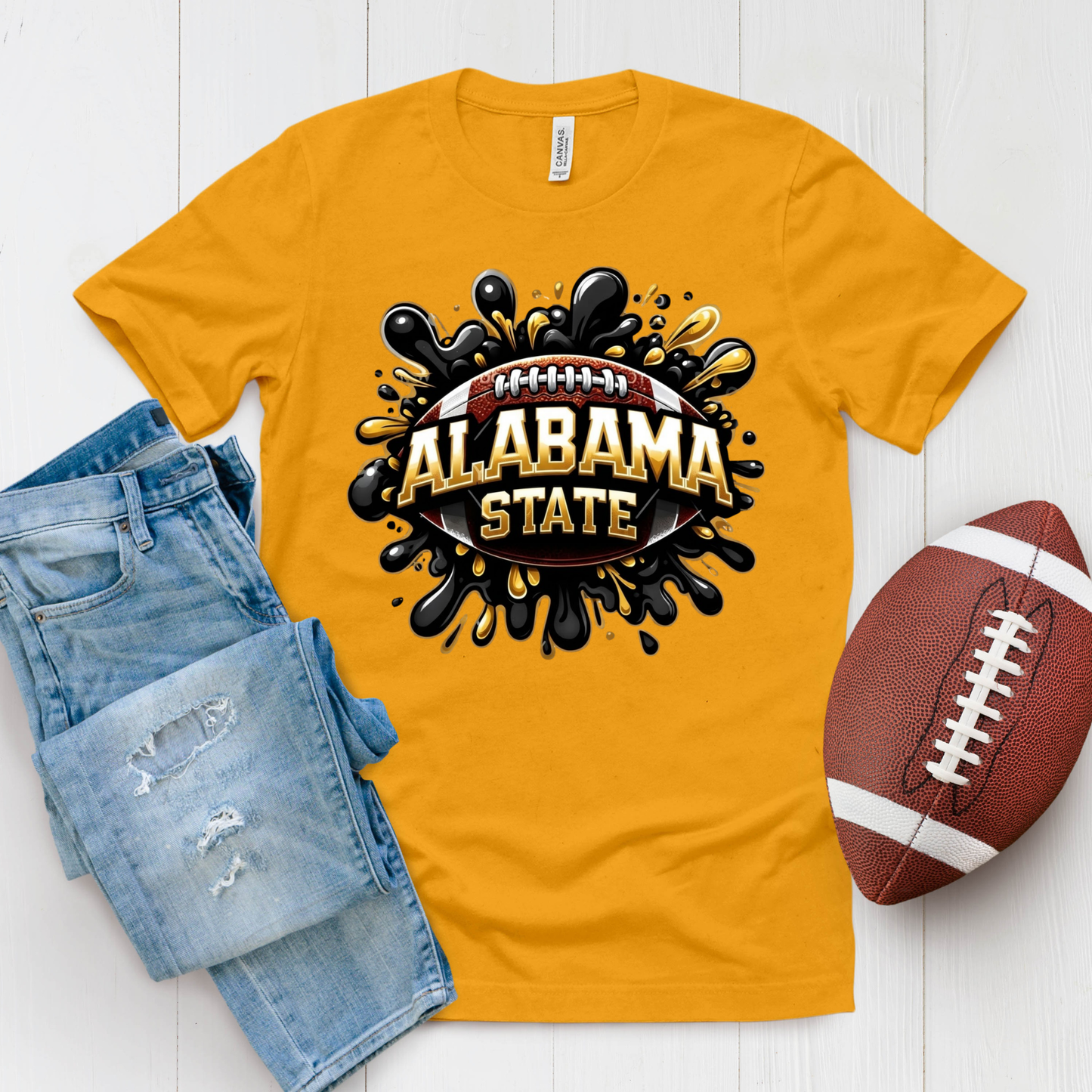 A gold short-sleeve t-shirt with a 'Hornet Nation Alabama State' graphic, styled in a flat lay with blue jeans and a brown football on a white wood background.