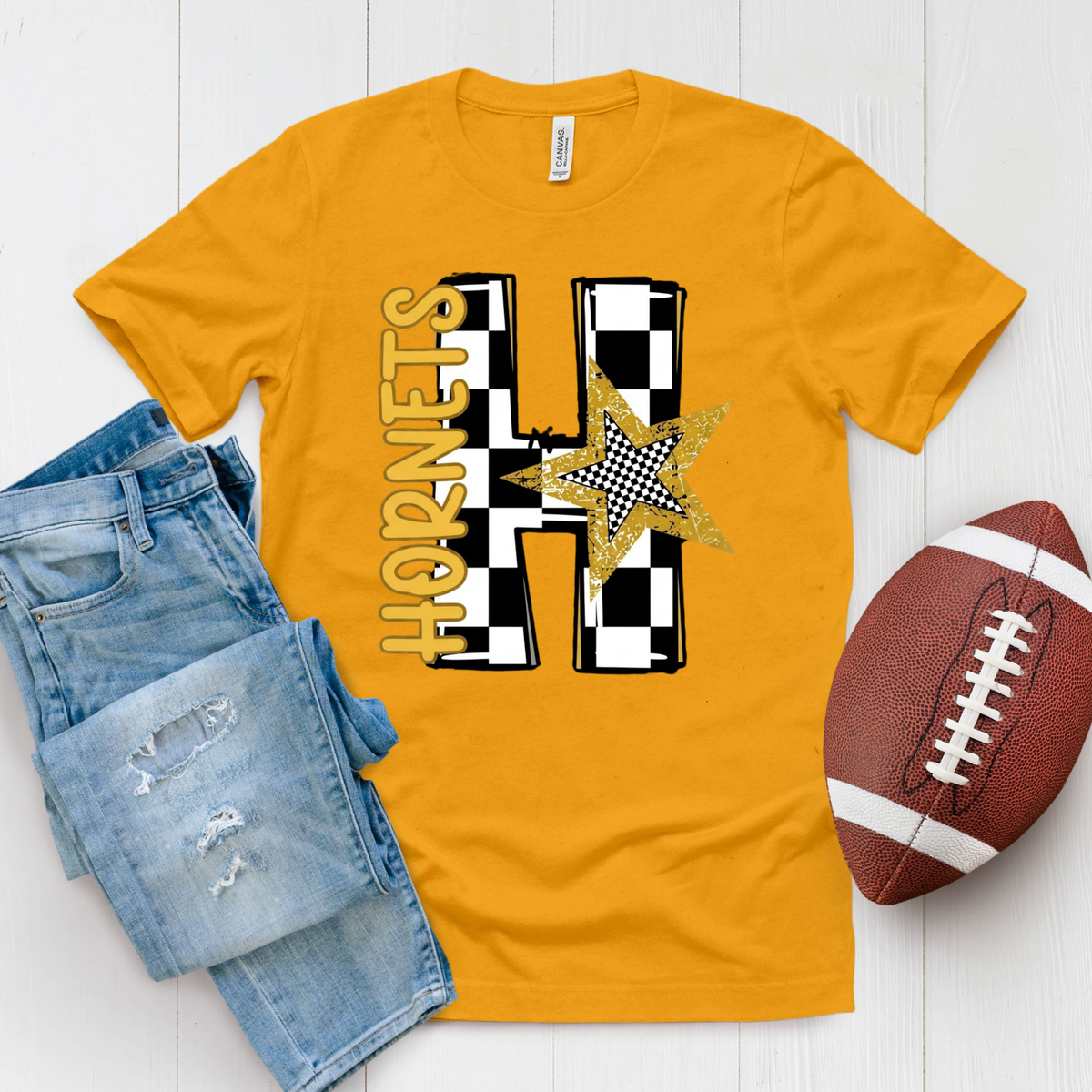 A gold short-sleeve t-shirt with a 'Hornet Nation Alabama State' graphic, styled in a flat lay with blue jeans and a brown football on a white wood background.