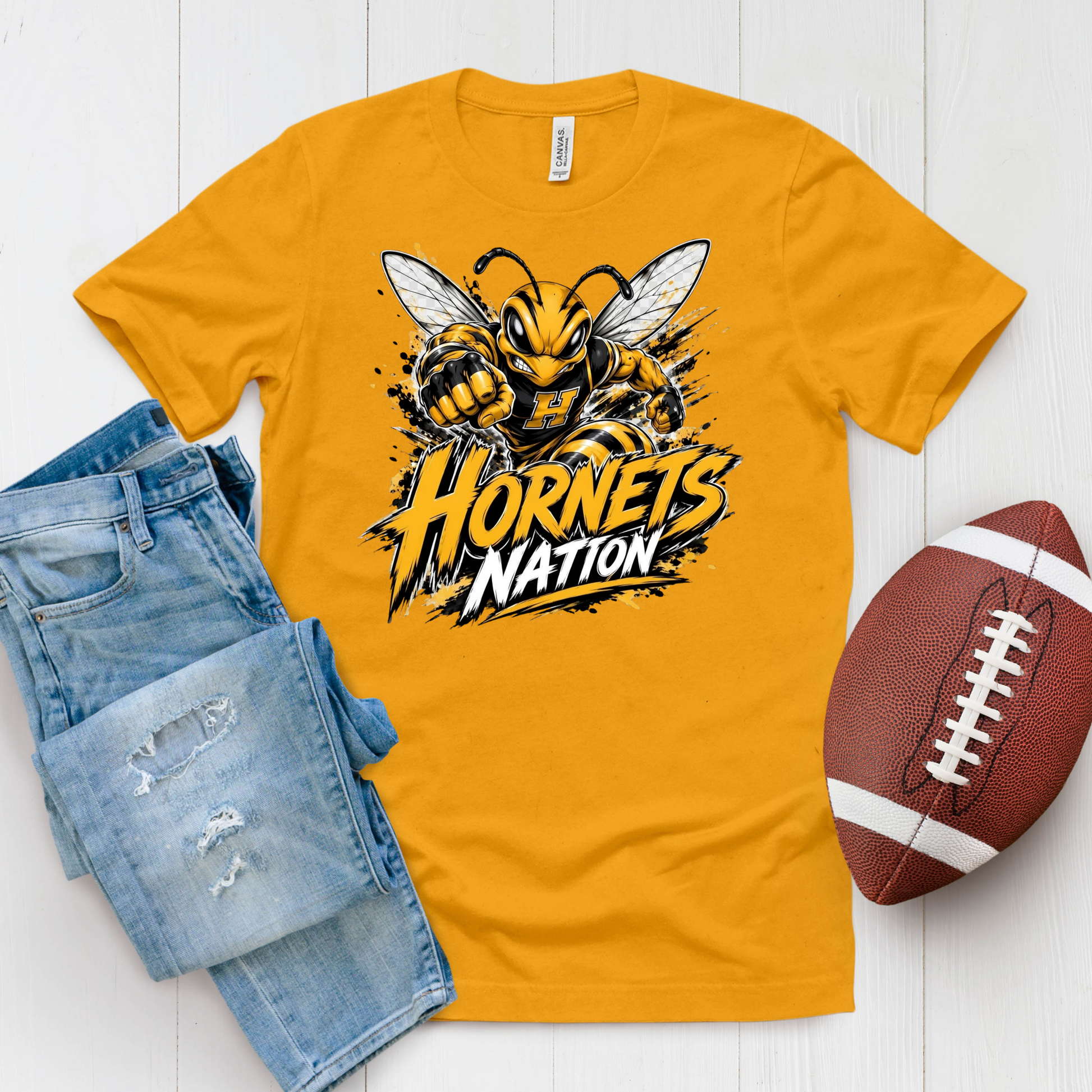 A gold crewneck t-shirt featuring a 'Hornets Nation' graphic with a hornet mascot, styled in a flat lay with blue jeans and a brown football on a white wood background.