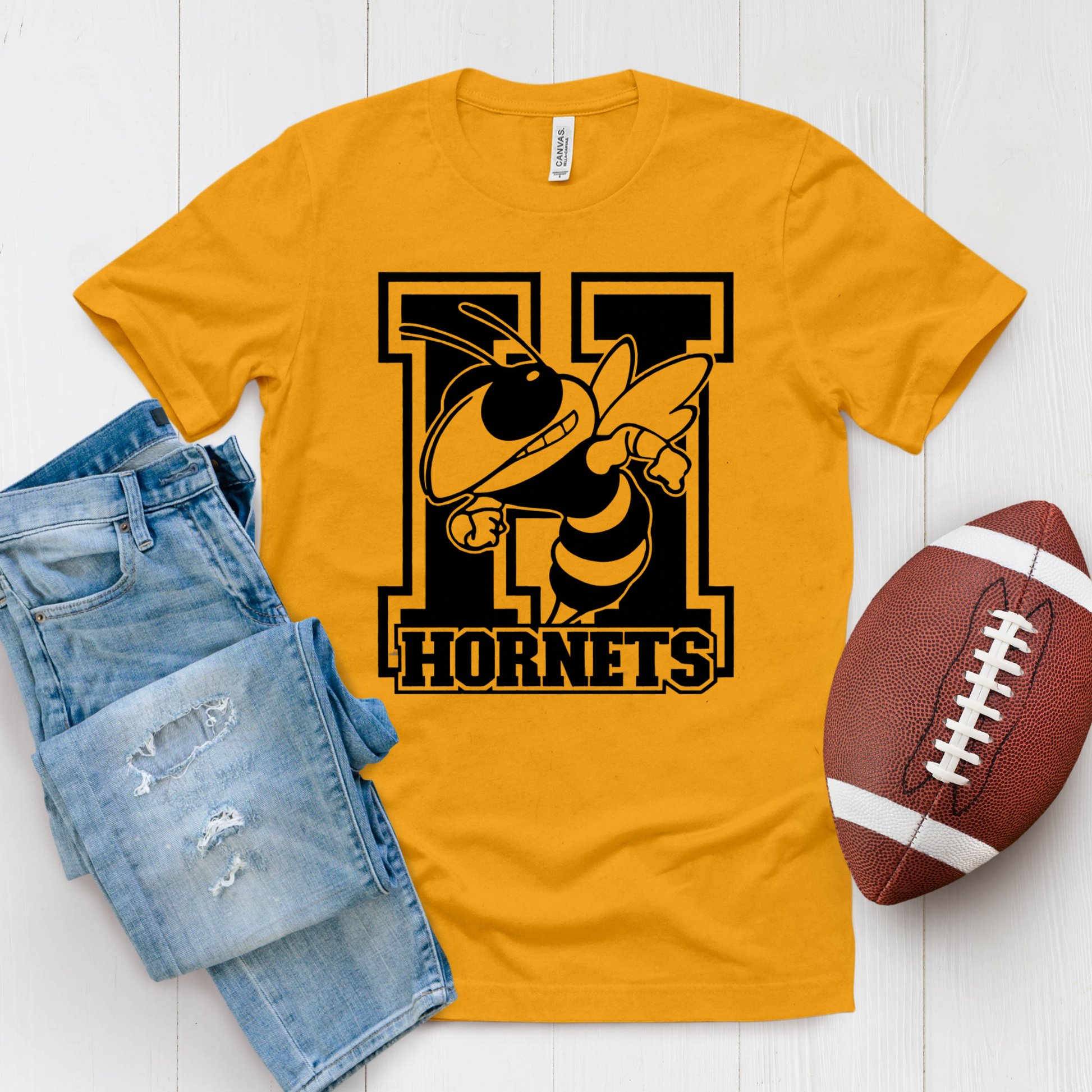 A gold crewneck t-shirt featuring a 'Hornets Nation' graphic with a hornet mascot, styled in a flat lay with blue jeans and a brown football on a white wood background.