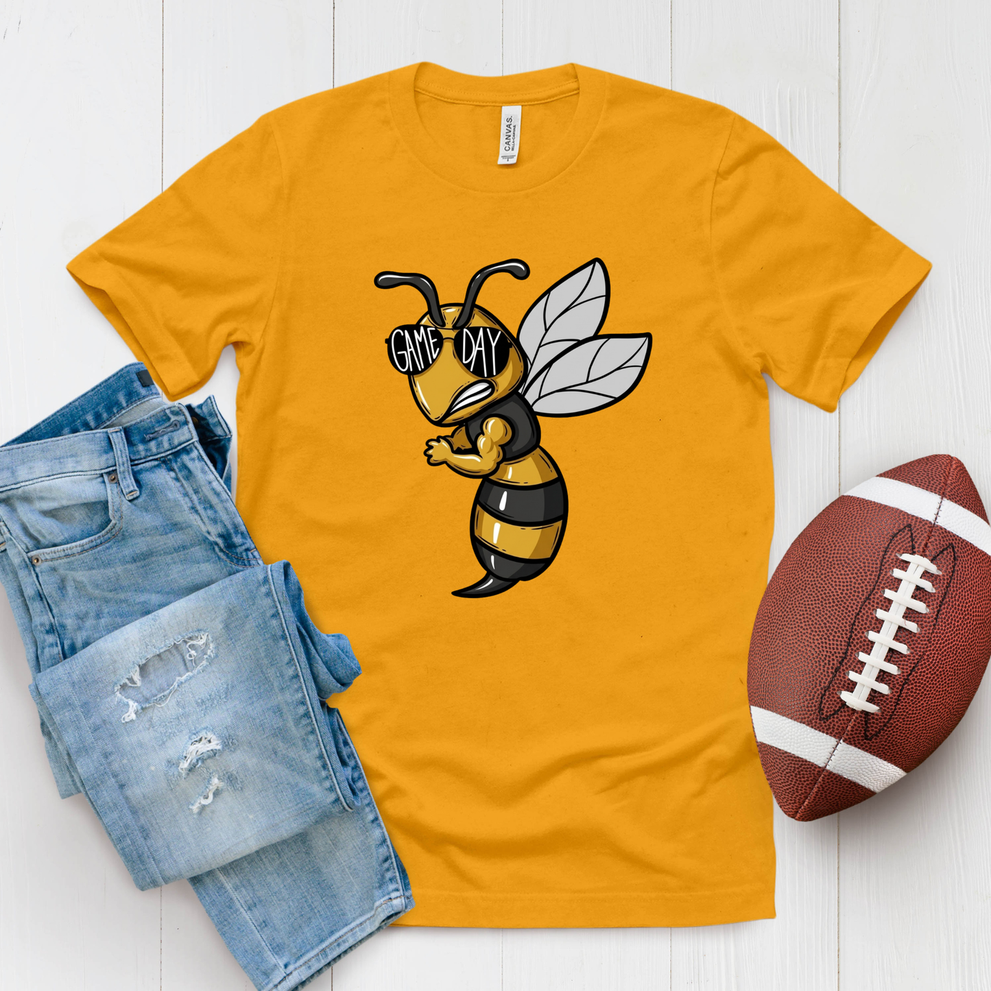 A gold crewneck t-shirt featuring a 'Hornets Nation' graphic with a hornet mascot, styled in a flat lay with blue jeans and a brown football on a white wood background.