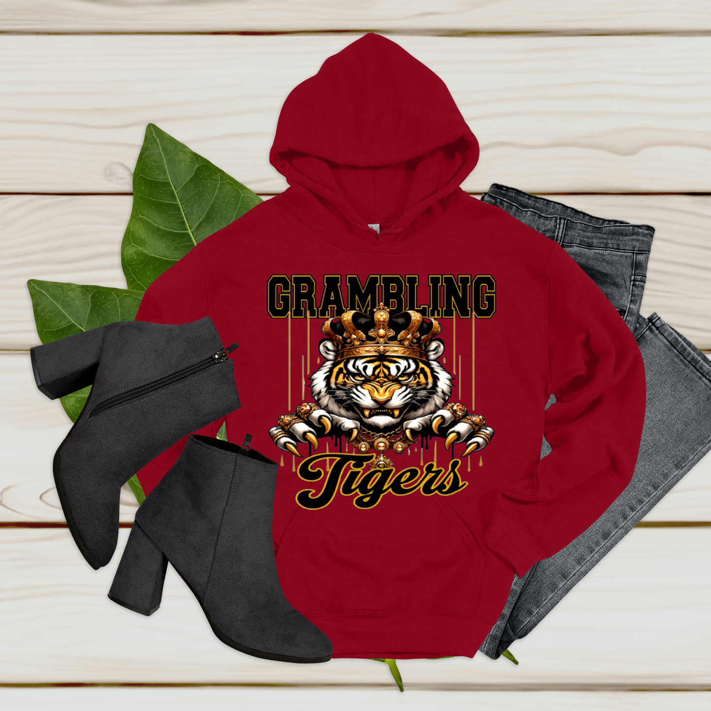 A red pullover hoodie with a tiger graphic and the words 'GRAMBLING Tigers' is styled in a flat lay on a light wood background, accompanied by black heeled ankle boots and folded gray jeans.
