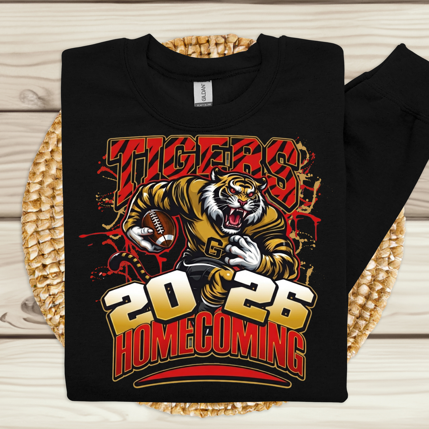 A black long-sleeve sweatshirt laid flat on a woven straw mat over a light wood surface. The front has a large graphic of a tiger football player with the words 'TIGERS 2026 HOMECOMING' in red, yellow, white, and black.