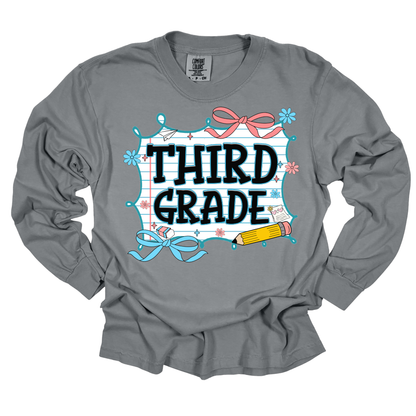 A gray long-sleeve t-shirt with a 'THIRD GRADE' school-themed graphic on the front, featuring a white lined notebook background, pink and blue bows, a yellow pencil, and small stars.