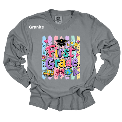 A gray long-sleeve t-shirt with a colorful 'First Grade' graphic on the front, featuring school-themed illustrations like a graduation cap, pencil, globe, and bus. The word 'Granite' is printed in white on the upper left chest.