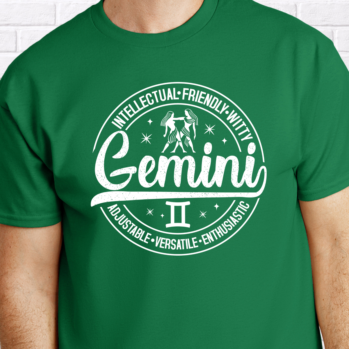 Forest green t-shirt laid flat, featuring a circular white Gemini zodiac graphic with the twins illustration, the word "Gemini," and trait keywords like intellectual, friendly, witty, enthusiastic, versatile, and adjustable.