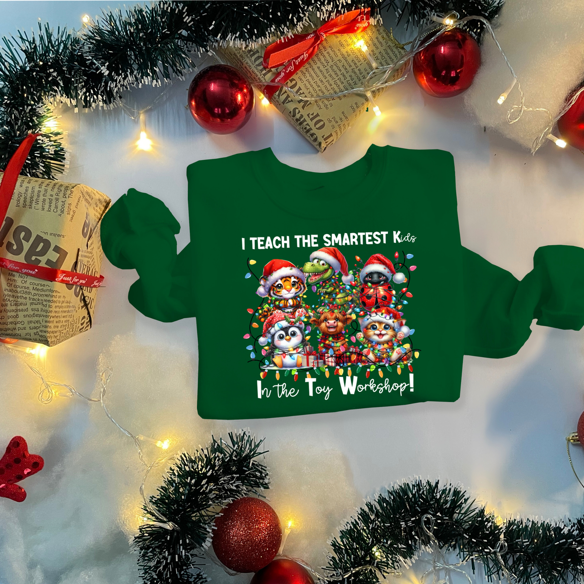A green long-sleeve sweatshirt with a colorful Christmas penguin graphic and the text 'I TEACH THE SMARTEST Kids In The Toy Workshop!' displayed in a festive flat-lay setting with holiday decorations.