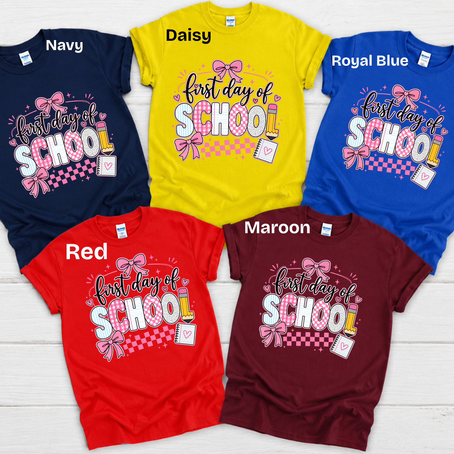 Five short-sleeve graphic T-shirts arranged on a white wood background, each labeled with a color name: Navy, Daisy yellow, Royal Blue, Red, and Maroon. The shirts all feature a colorful 'FIRST Grade' text graphic on the chest.