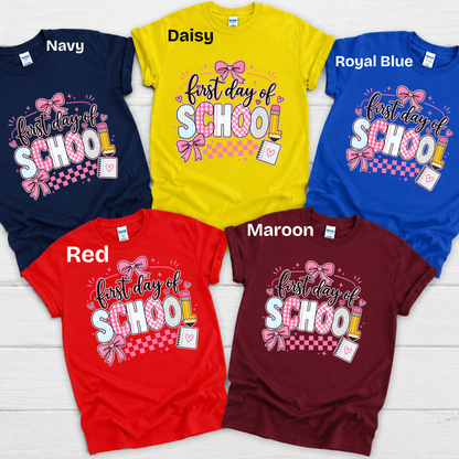 Five short-sleeve graphic T-shirts arranged on a white wood background, each labeled with a color name: Navy, Daisy yellow, Royal Blue, Red, and Maroon. The shirts all feature a colorful 'FIRST Grade' text graphic on the chest.
